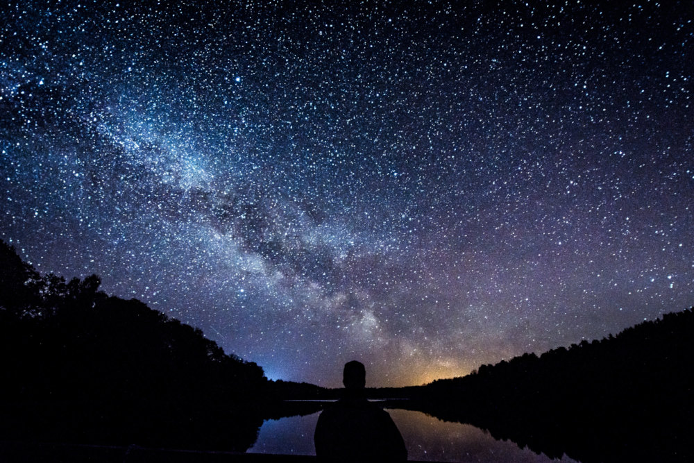 starry sky, faint light behind forest - photo by mindaugas vitkus